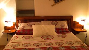 Een slaapkamer met een houten hoofdbord, een bed met beddengoed met hartjes, twee bedlampjes en een plank met foto's.