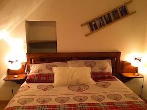 A bedroom with a wooden headboard, a bed with heart-patterned bedding, two bedside lamps, and a shelf with pictures.
