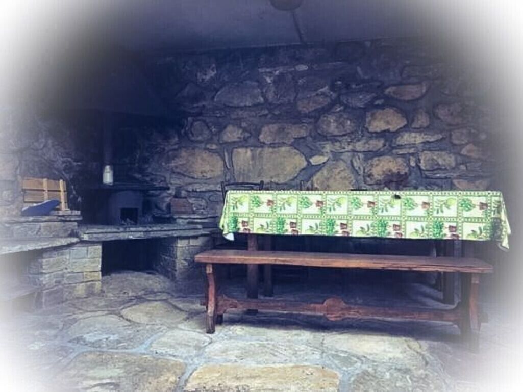 Une cheminée en pierre avec un banc devant, une table recouverte d’une nappe à motifs et un mur en pierre rustique.