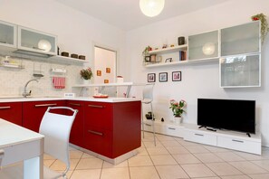 A modern kitchen with red cabinets, a white countertop, a built-in TV, and a dining area with a white chair.