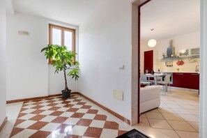 A room with a checkered floor, a potted plant, a window, and a view into a kitchen area.