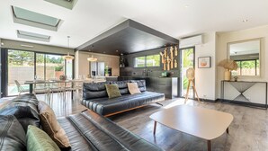 A modern living room with a black leather sofa, a white coffee table, and a dining area with a glass table and chairs. - Modern Villa with Private Pool (Saint-Aignan-Grandlieu)