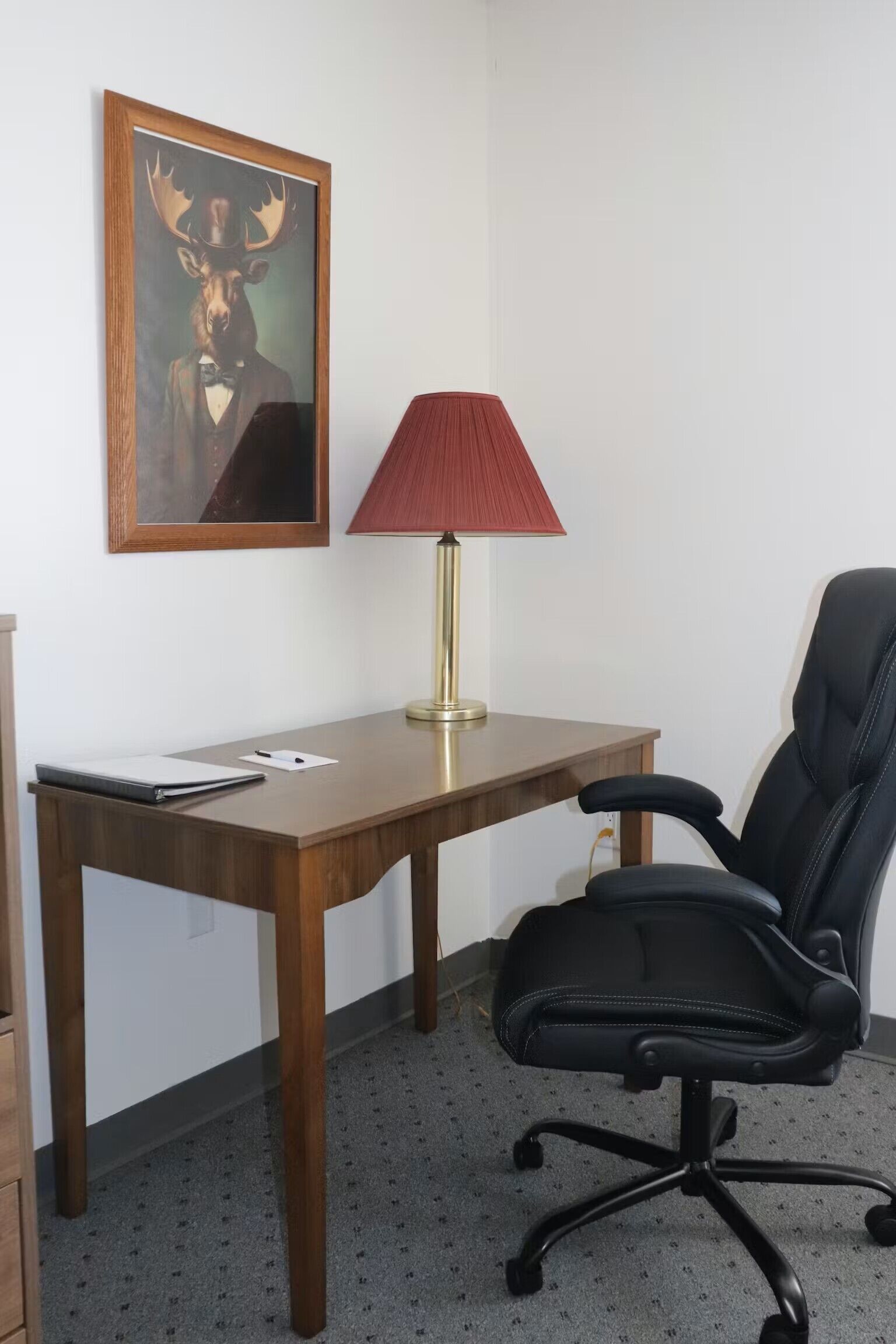 Un bureau en bois, une lampe rouge, un fauteuil de bureau noir et, au mur, un tableau représentant un élan.