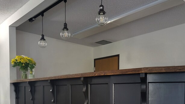 A reception desk with a dark wood finish, three hanging pendant lights, and a vase with yellow flowers on the left. - Piper's Inn (Clearbrook)