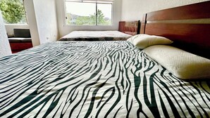 A bedroom with a zebra-patterned bedspread, a wooden headboard, a window with blinds, and a view of greenery outside.