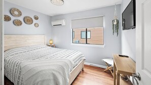 A bedroom with a wooden bed, a bedside table, a hanging plant, a TV, and a window with blinds. - Modern, renovated, centrally located condo with self check in and parking (Ocean City)