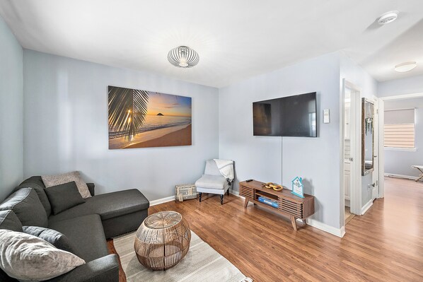 Un salon moderne avec un canapé d’angle, un téléviseur à écran plat, un tableau représentant une plage et une table basse en bois.