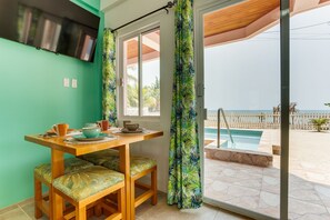 A dining area with a table and chairs, a television mounted on the wall, and a view of a swimming pool outside.