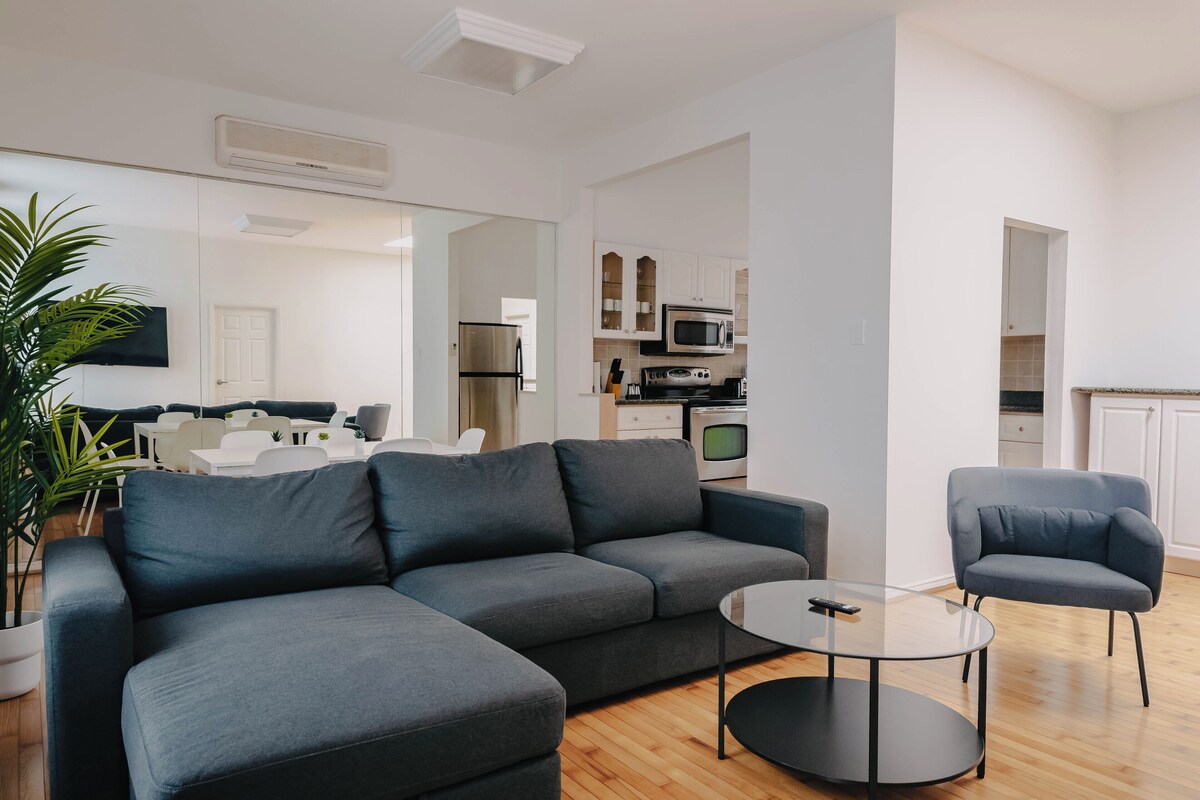 A modern living room with a grey sofa, a round coffee table, and a blue armchair. A dining area with white furniture is visible in the background.