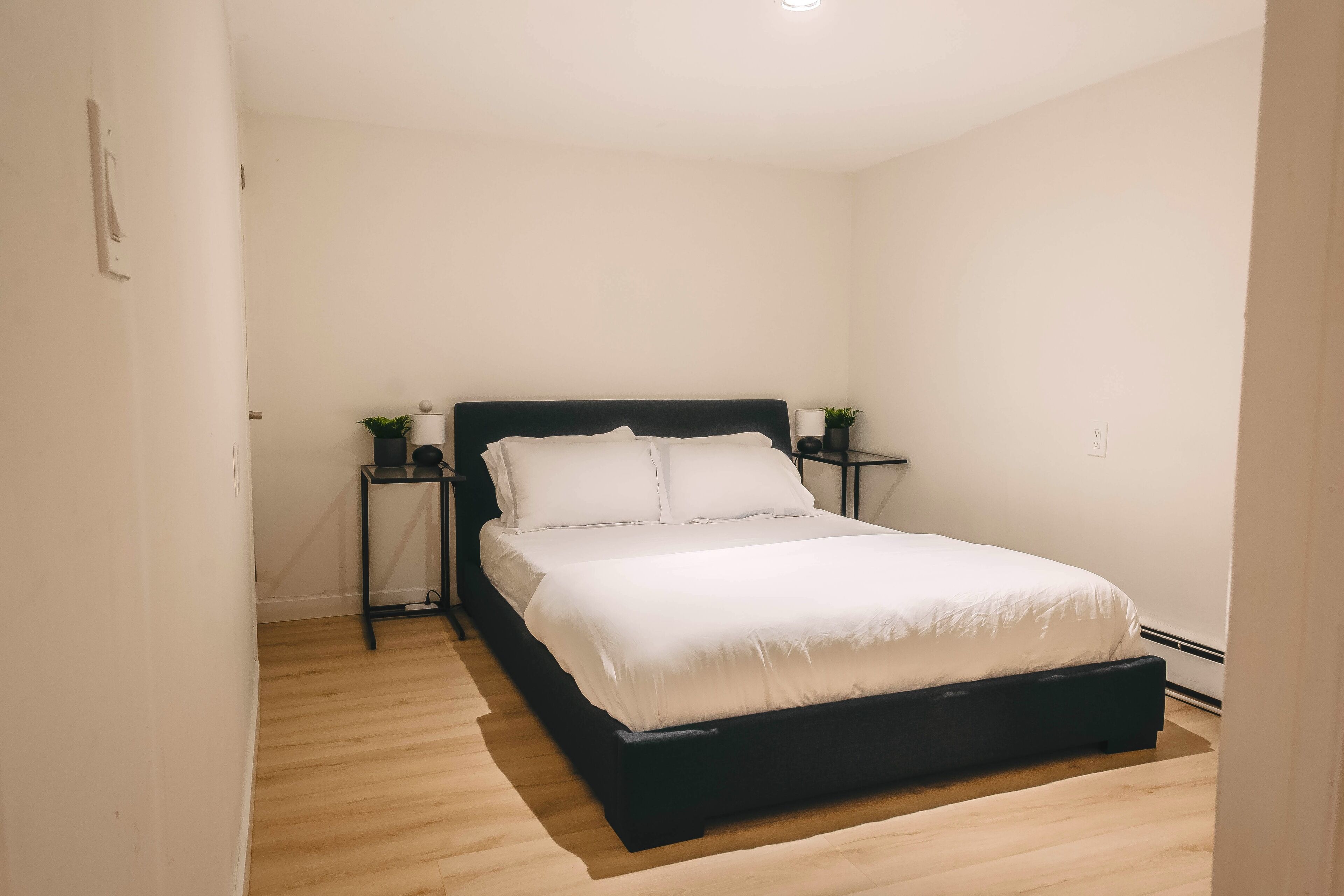 Une chambre à coucher avec un cadre de lit noir, du linge de lit blanc et deux tables de chevet ornées de lampes et de petites plantes.