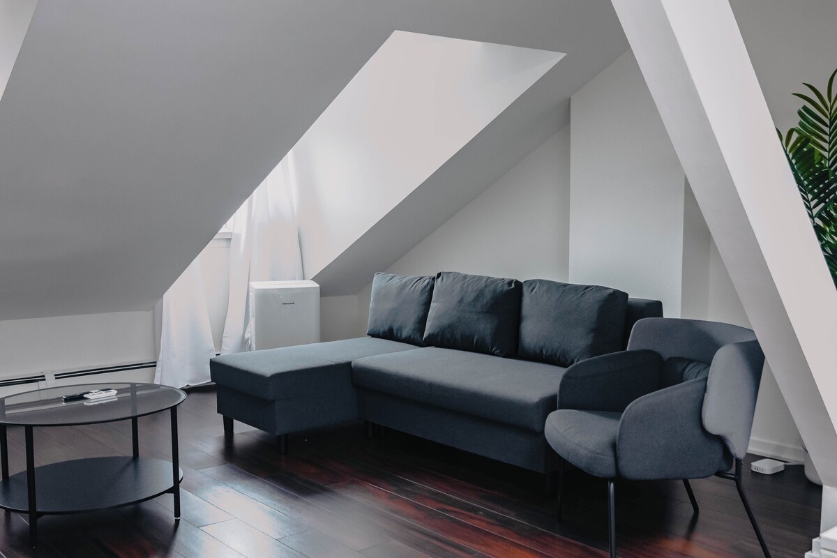 A modern loft bedroom with a sloped ceiling, a gray sectional sofa, a round coffee table, and a potted plant.