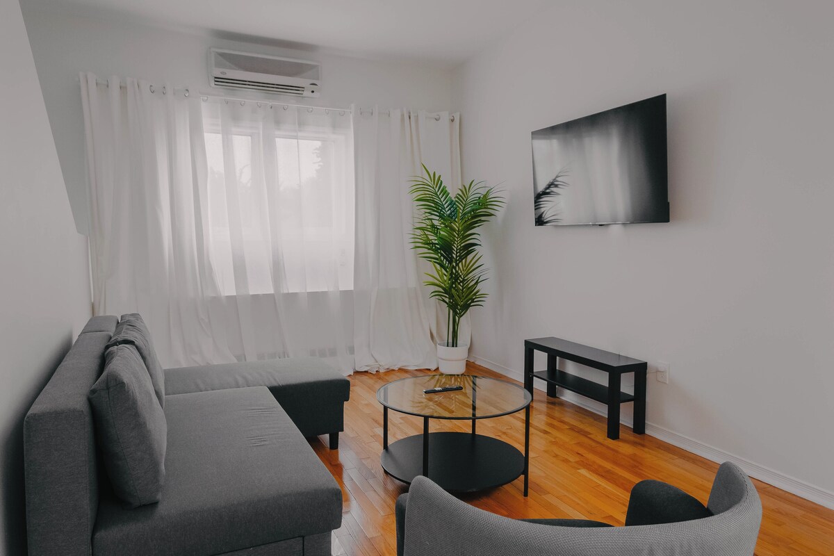 A modern living room with a grey sofa, a round glass coffee table, a black side table, a potted plant, and a mounted TV on the wall.