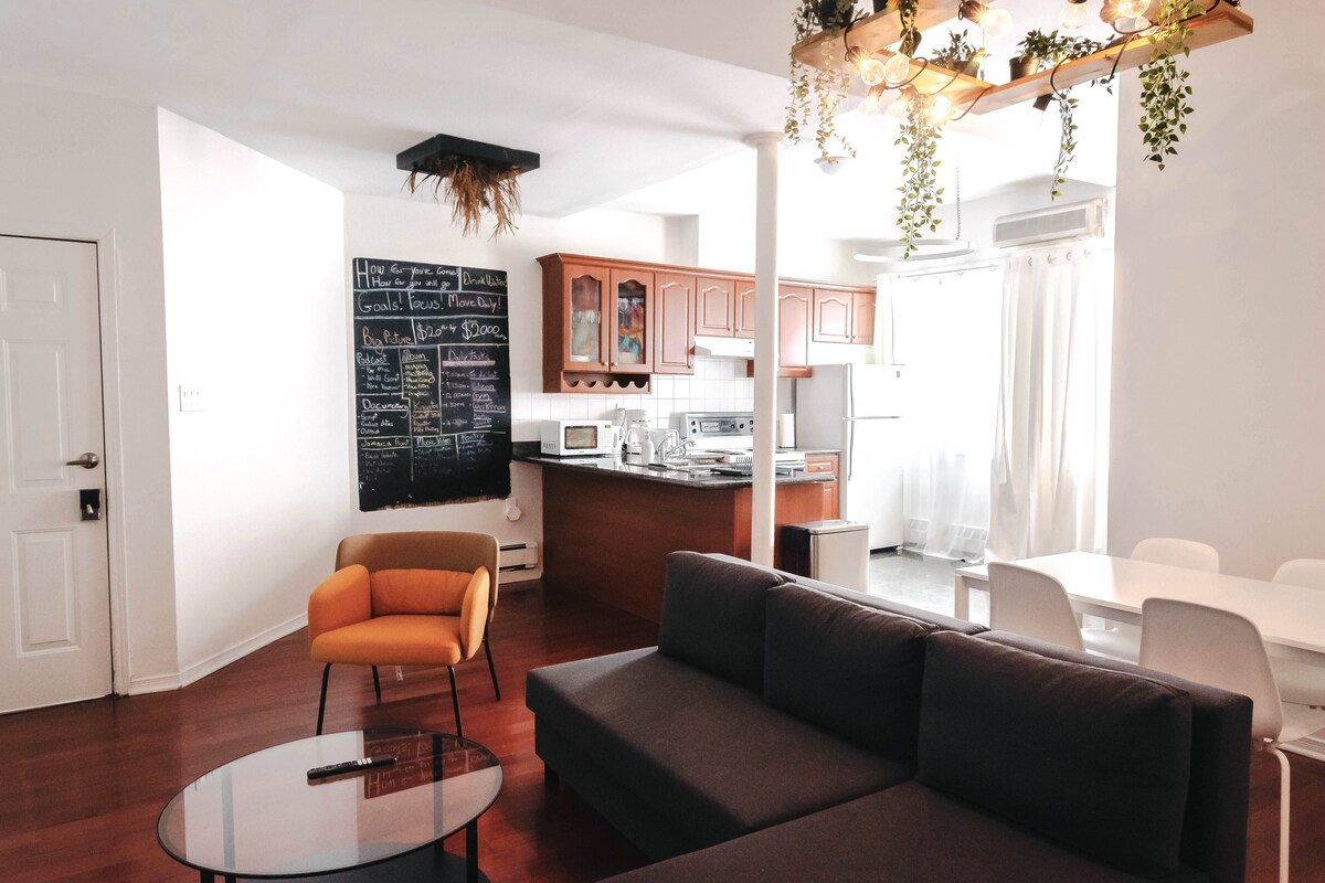 A modern living room with a sofa, a glass coffee table, and an orange chair. A chalkboard with goals and a refrigerator are visible in the background.