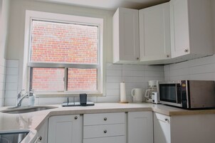 Une cuisine avec des meubles blancs, une fenêtre donnant sur un mur de briques, un four à micro-ondes et une machine à café.