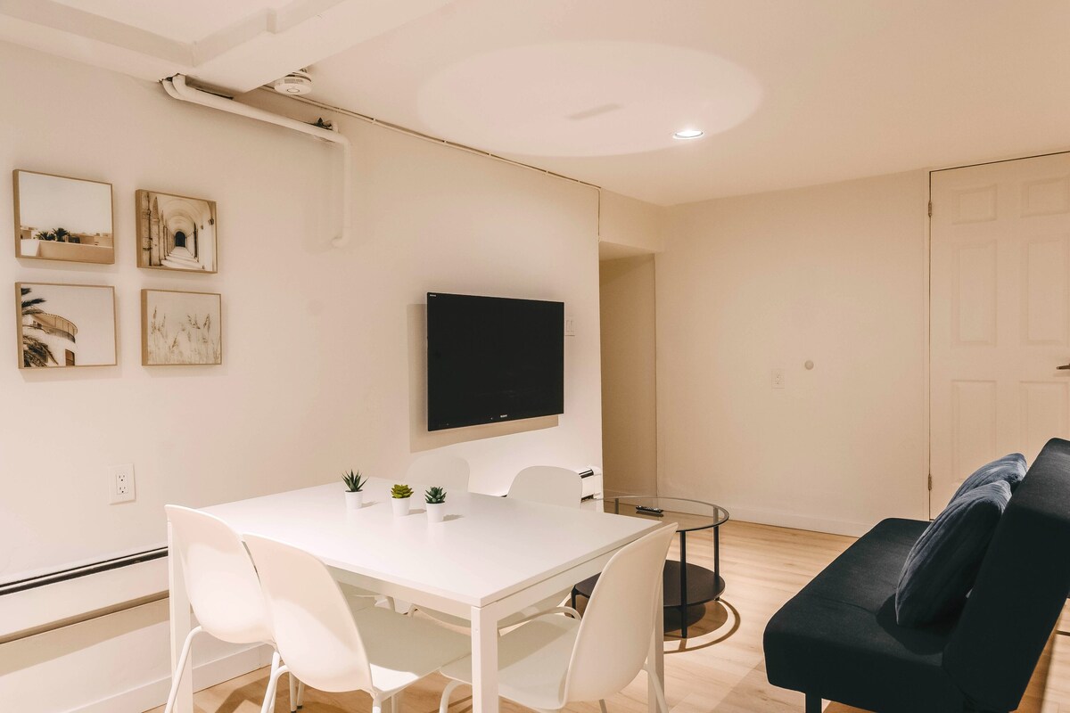 A modern living room with a white table, white chairs, a black sofa, and a flat-screen TV mounted on the wall.