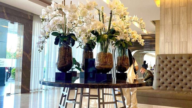 Un hall d’entrée avec une table ronde en verre, entourée de vases contenant des orchidées blanches, un éclairage moderne suspendu et un canapé capitonné.