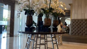 Un hall d’entrée avec une table ronde en verre, entourée d’orchidées blanches, agrémenté de luminaires suspendus modernes et d’un canapé capitonné.