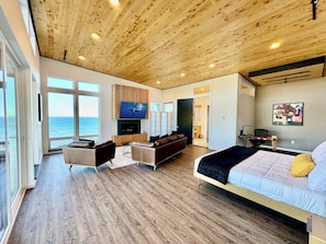 Ein modernes Schlafzimmer mit einem großen Bett, einem an einer Holzvertäfelung montierten Fernseher, einer Sitzecke mit Sofa und Sesseln sowie Meerblick durch große Fenster.