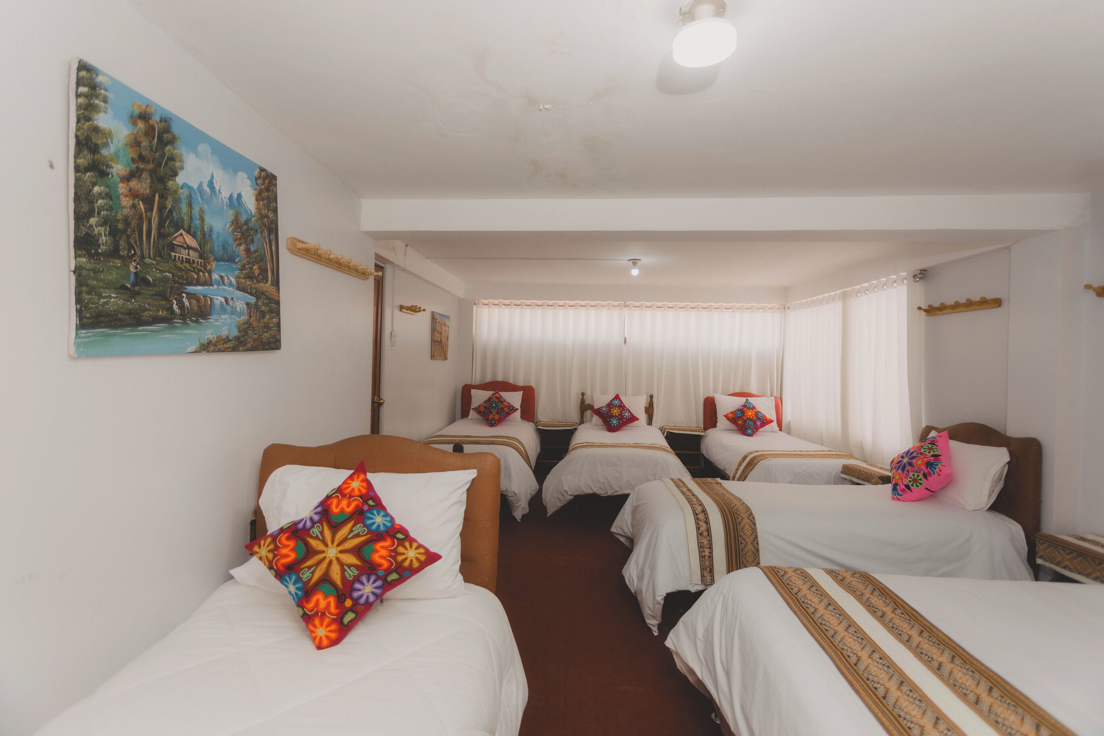 Une chambre d’hôtel avec quatre lits simples, du linge de lit blanc, des oreillers colorés, un tableau représentant un paysage accroché au mur et une grande fenêtre avec des rideaux.