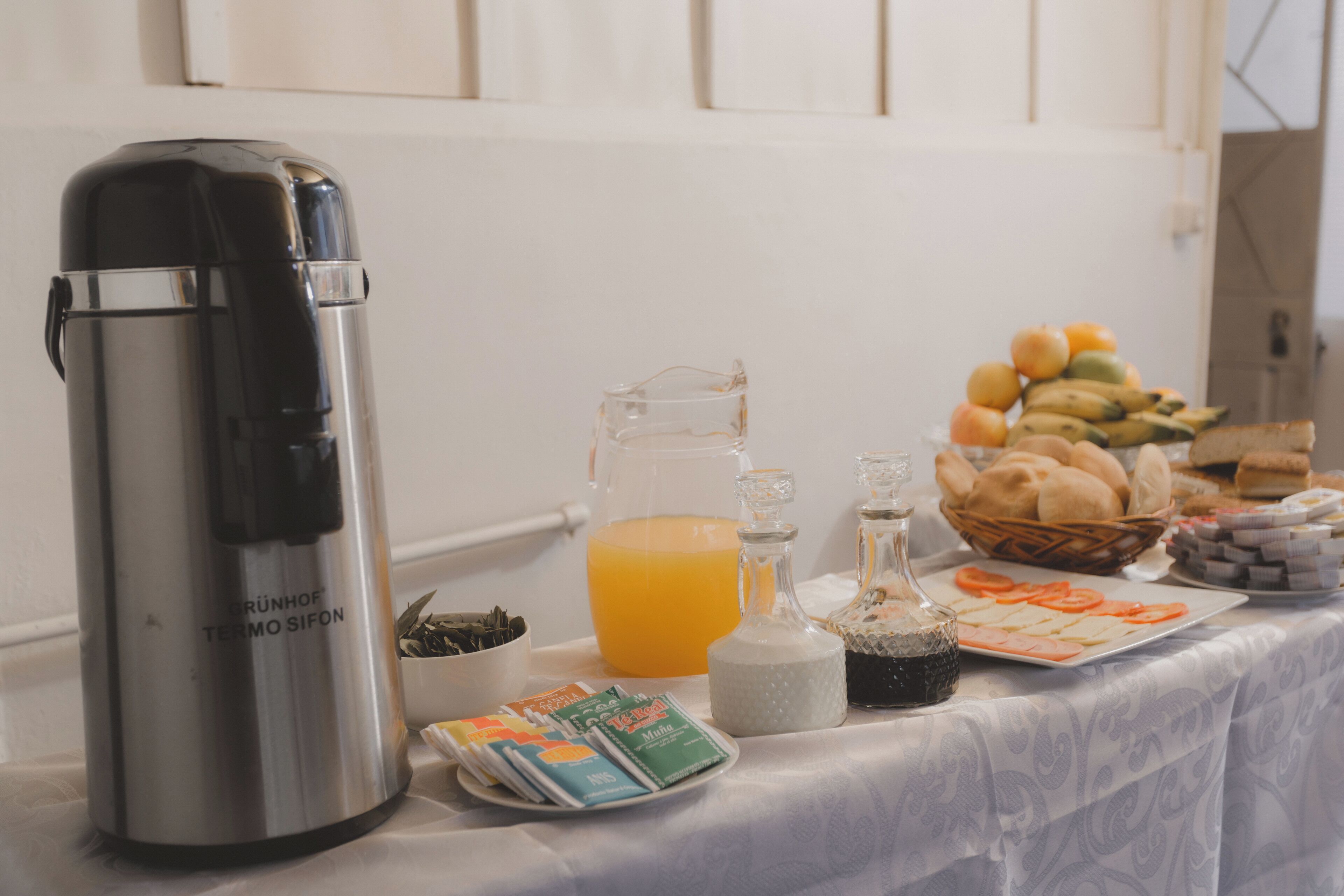 A breakfast table with a coffee maker, juice, condiments, bread, and fruit.