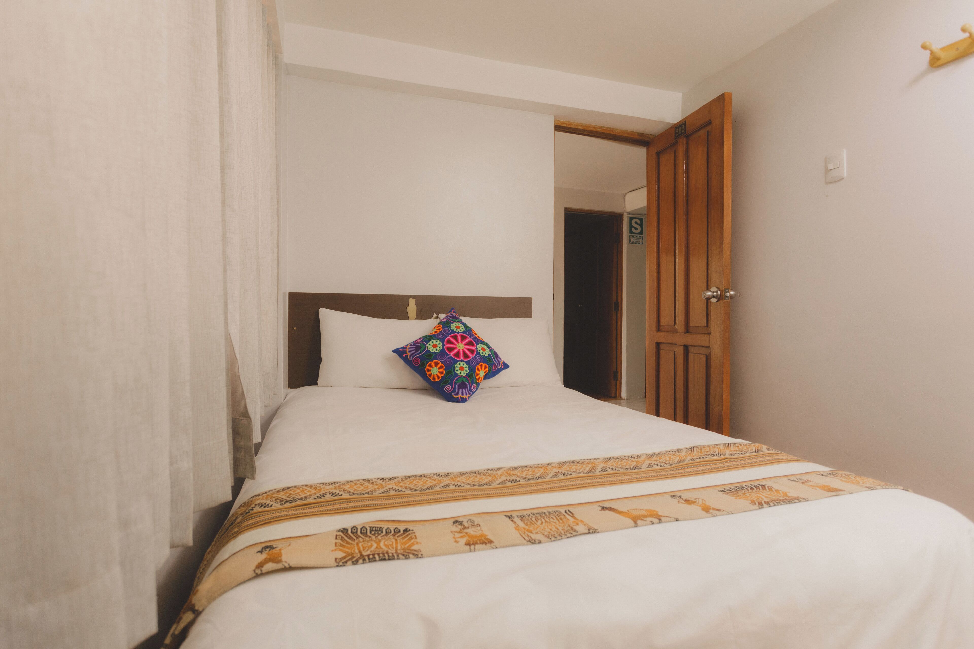 A neatly made bed with a patterned bedspread and a colorful pillow, a wooden door to the right, and a white wall.
