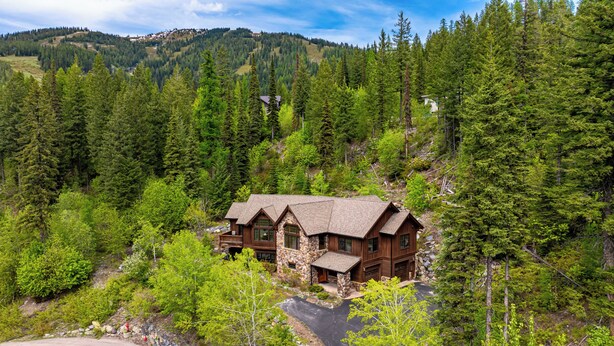 A log cabin with a stone foundation, surrounded by dense forest and hills in the background.