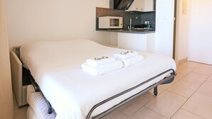 Een net opgemaakt bed met witte lakens en handdoeken, een kleine kitchenette met een magnetron en een badkamer met een douche.