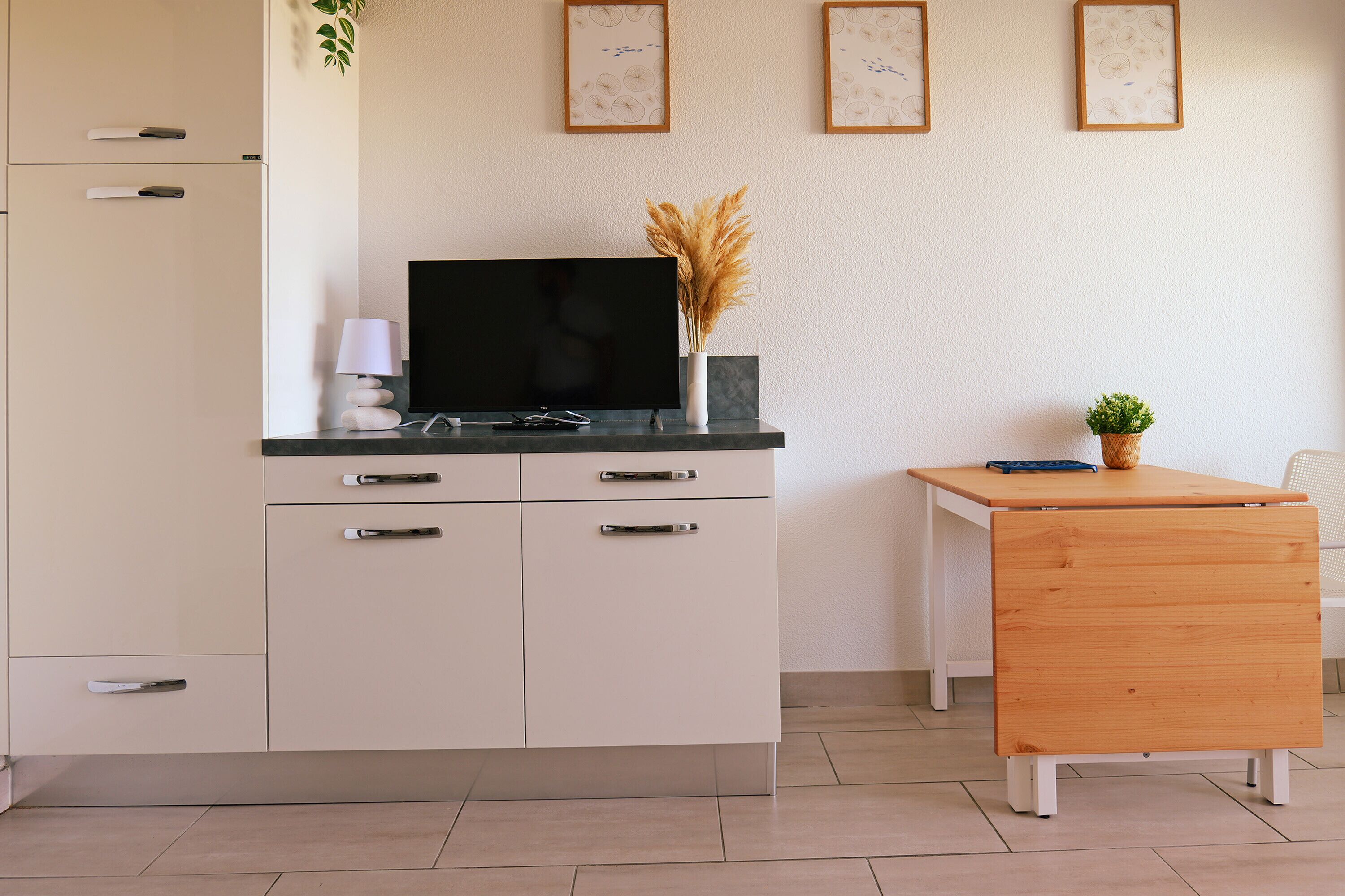 A modern kitchen with a flat-screen TV, white cabinets, a wooden table, and decorative plants.