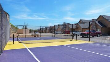 A tennis court with a net, surrounded by a fence, and vehicles parked nearby.
