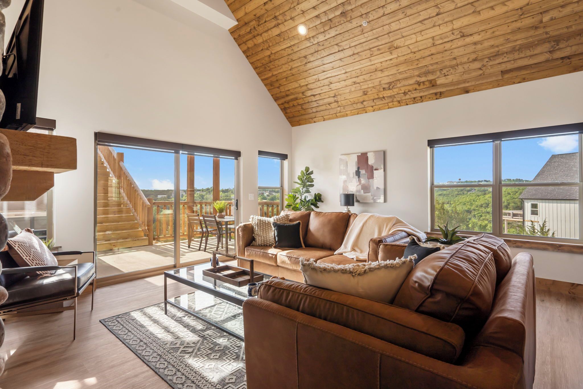 A spacious living room with a high ceiling, a large brown leather sofa, a glass coffee table, and a view of a wooden staircase leading to a balcony.