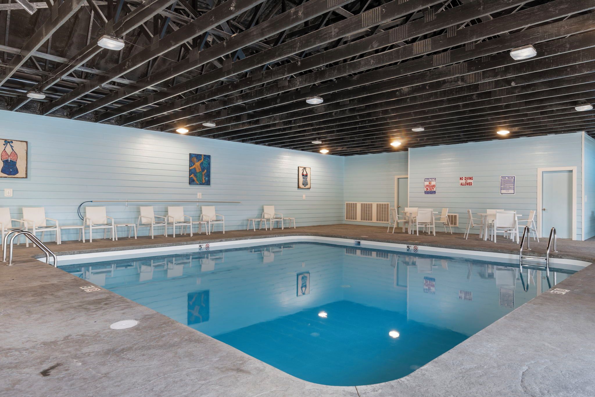 An indoor swimming pool with a blue floor, surrounded by white chairs and a lifeguard station.