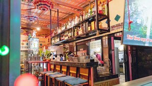A bar area with multiple shelves of bottles, a bar counter, and seating with cushions. - Touch the Wind (Sap Yai)