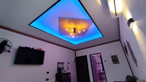 A room with a ceiling light fixture, a flat-screen TV mounted on the wall, and a door leading to another room. - Touch the Wind (Sap Yai)