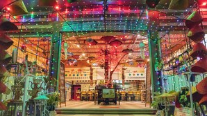 A brightly lit indoor space with a central tree, tables, chairs, and a sign that reads "WELCOME". - Touch the Wind (Sap Yai)