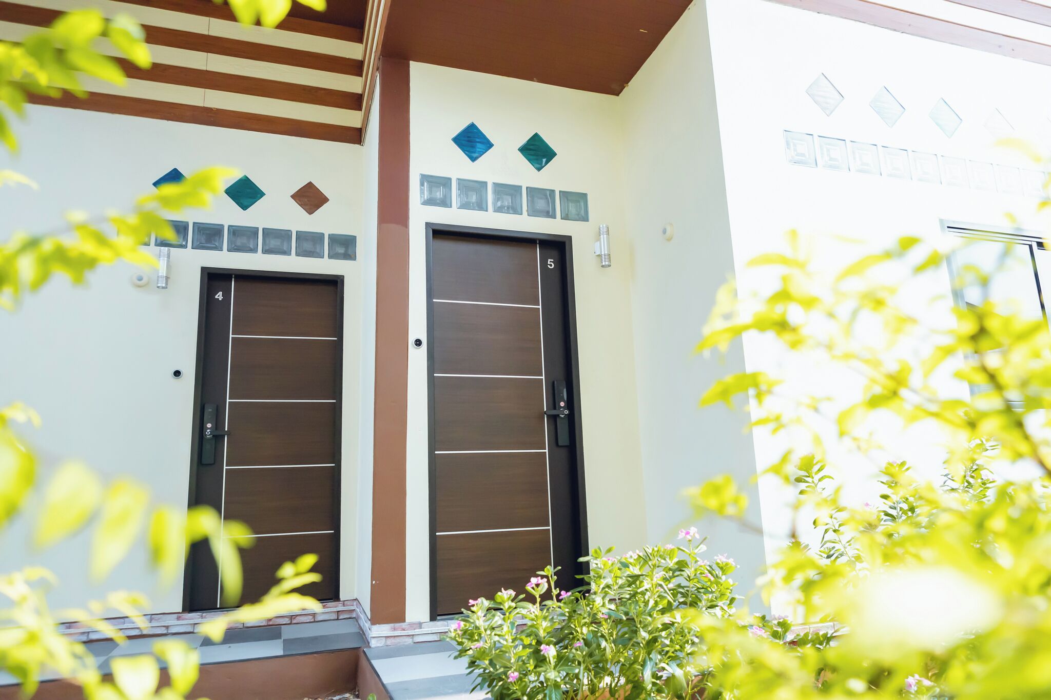 Une entrée d’immeuble avec deux portes, un mur recouvert de carreaux orné d’éléments décoratifs et un petit jardin avec des plantes.