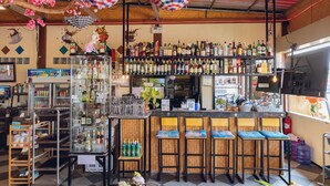 A bar area with shelves of liquor bottles, a counter with stools, and hanging decorations. - Touch the Wind (Sap Yai)