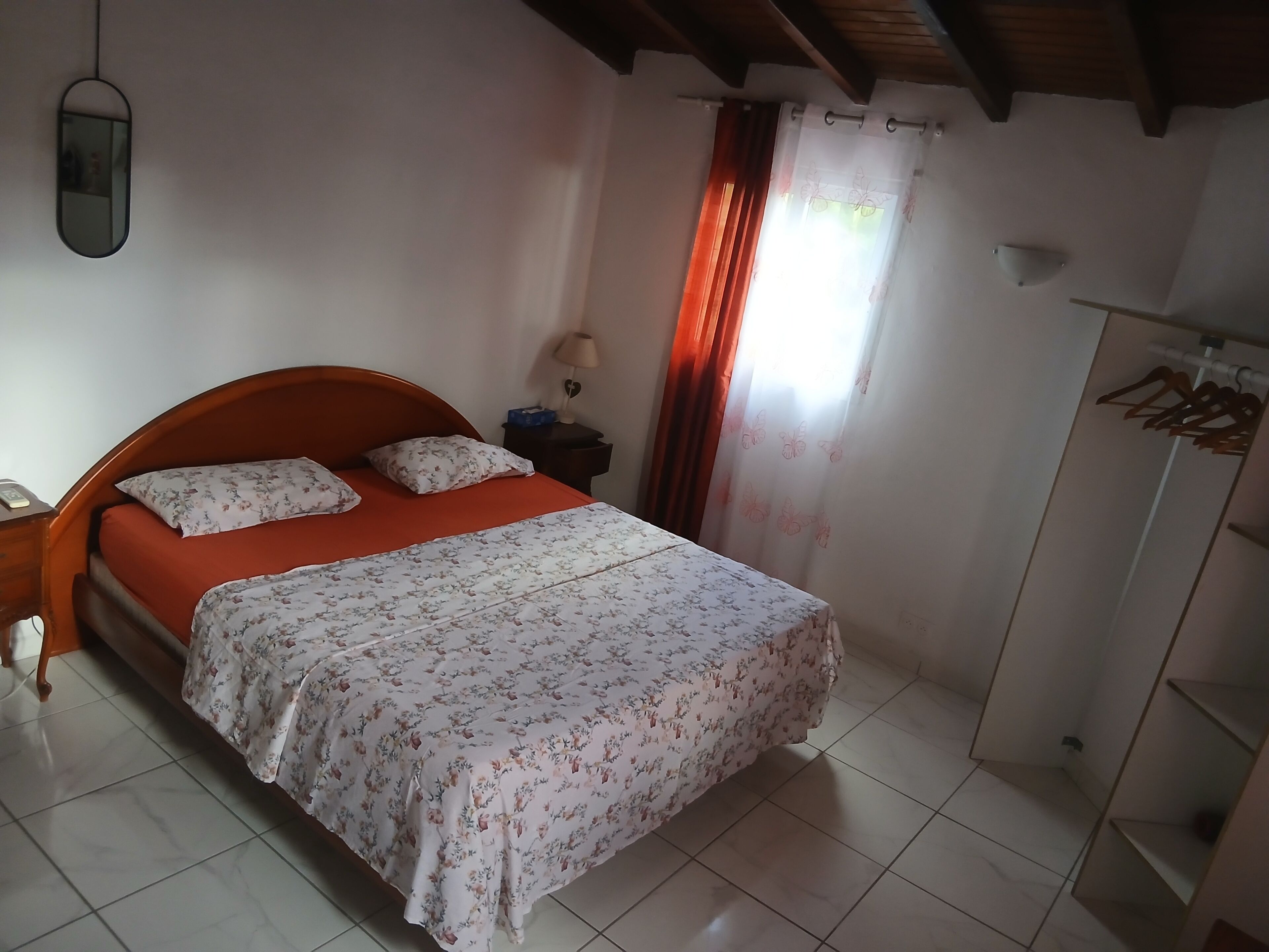 A bedroom with a bed, a nightstand, a mirror, a wardrobe, and a window with curtains.