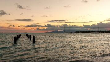 A sunset over a calm sea with wooden posts in the foreground and boats in the background.