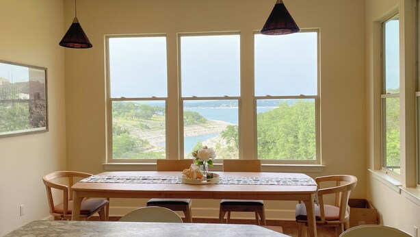 Ein Essbereich mit einem großen Tisch, Stühlen und Blick auf eine natürliche Landschaft durch drei Fenster.