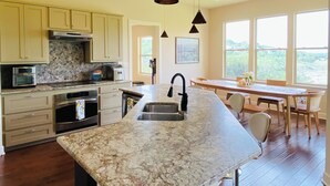 Eine moderne Küche mit Granit Arbeitsplatten, Edelstahlgeräten und einer großen Kochinsel mit Spüle. Durch ein großes Fenster ist ein Essbereich mit Tisch und Stühlen zu sehen.