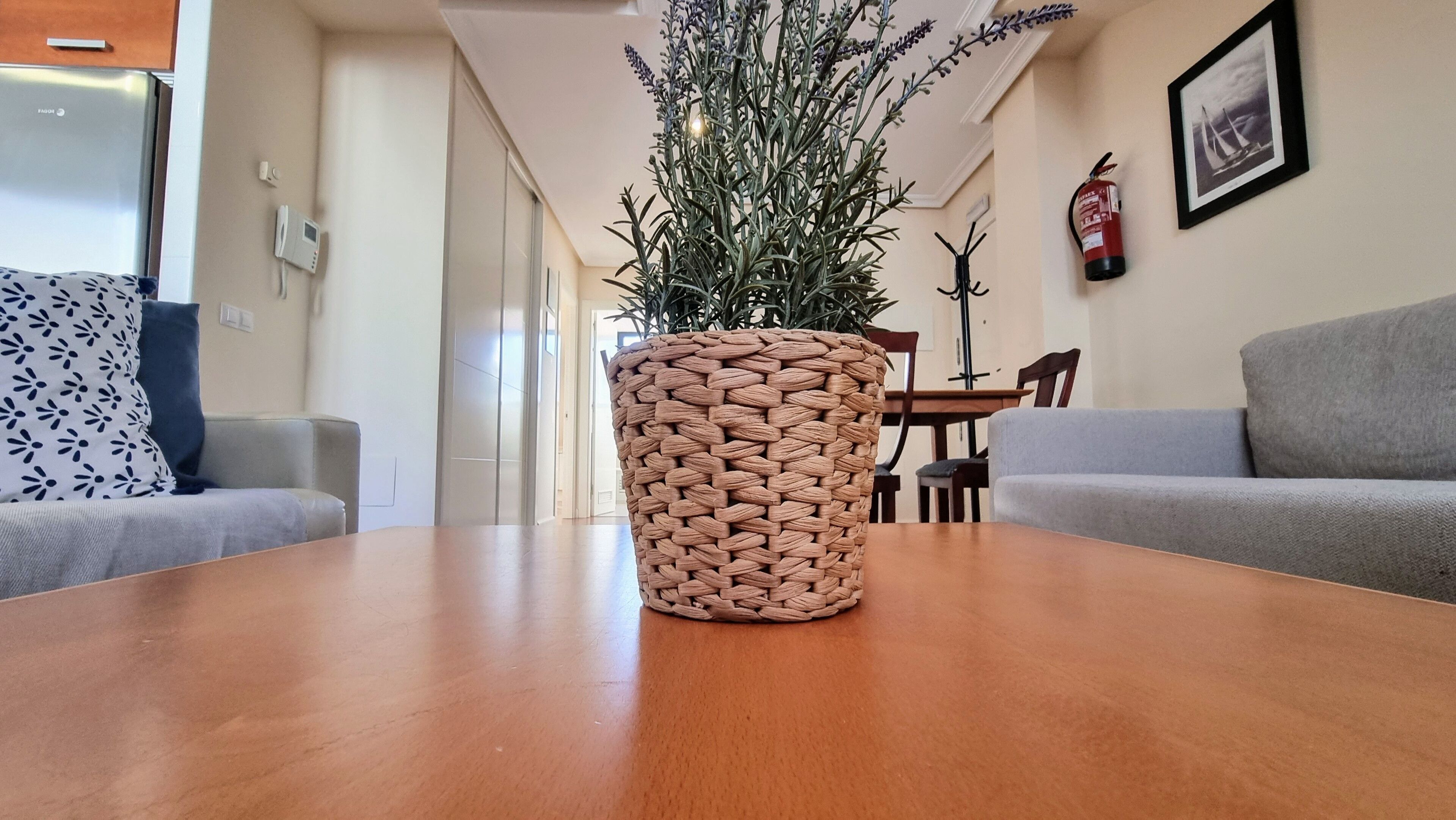 Una mesa de madera con una maceta tejida en el centro, flanqueada por dos sofás grises. En el fondo, se ve una mesa de comedor y sillas.