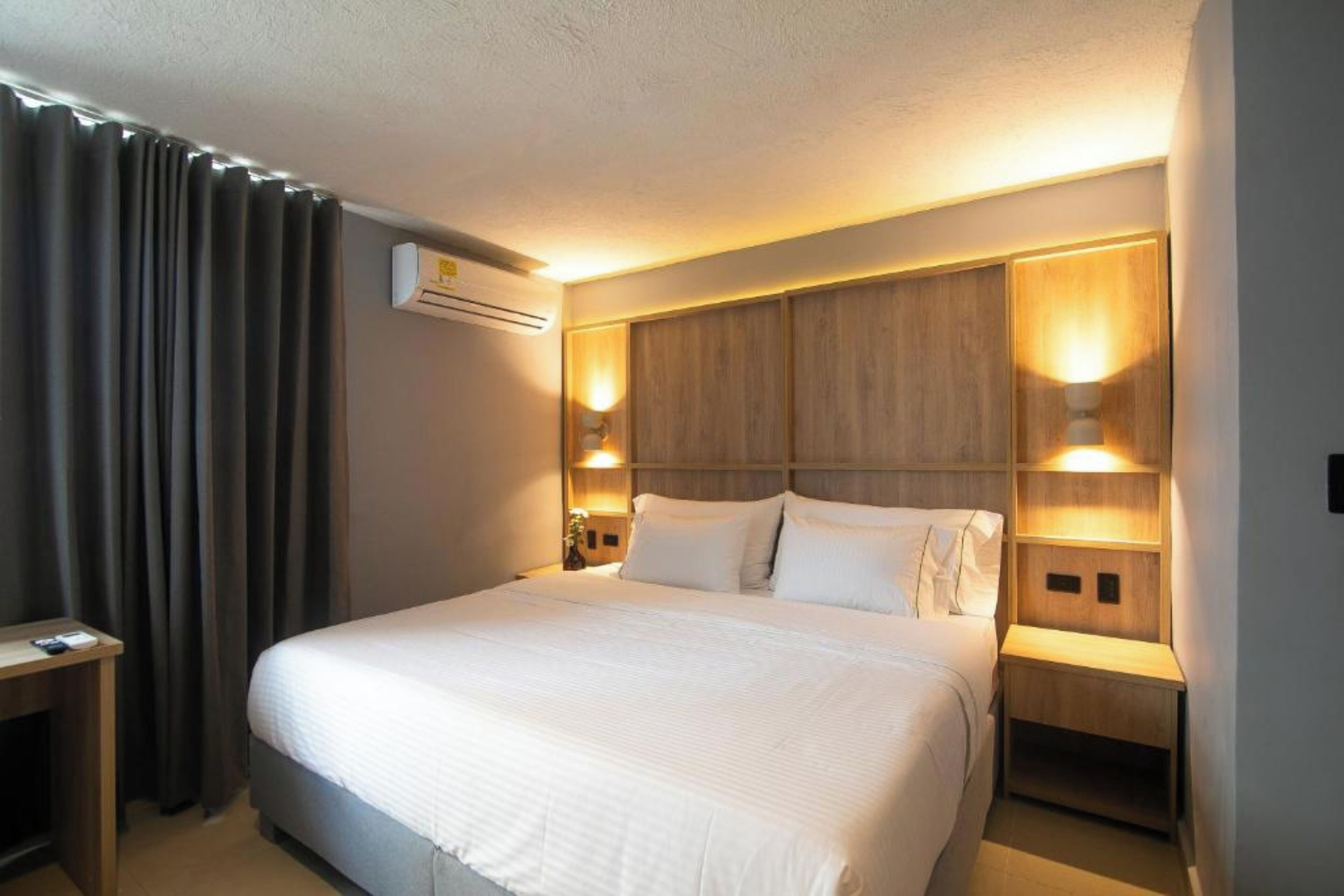 A hotel room with a large bed, two bedside tables, a desk, and a wall-mounted air conditioner.