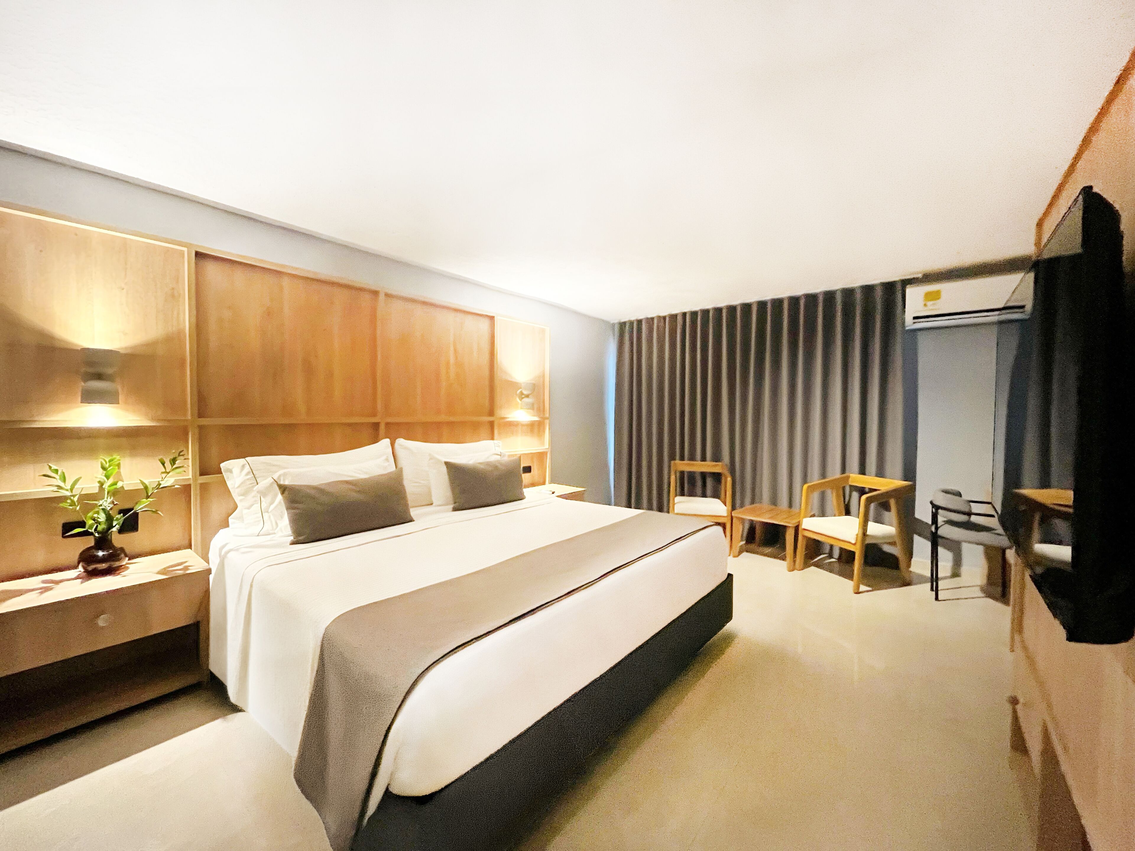 A hotel room with a bed, bedside tables, a desk, chairs, and an air conditioning unit.
