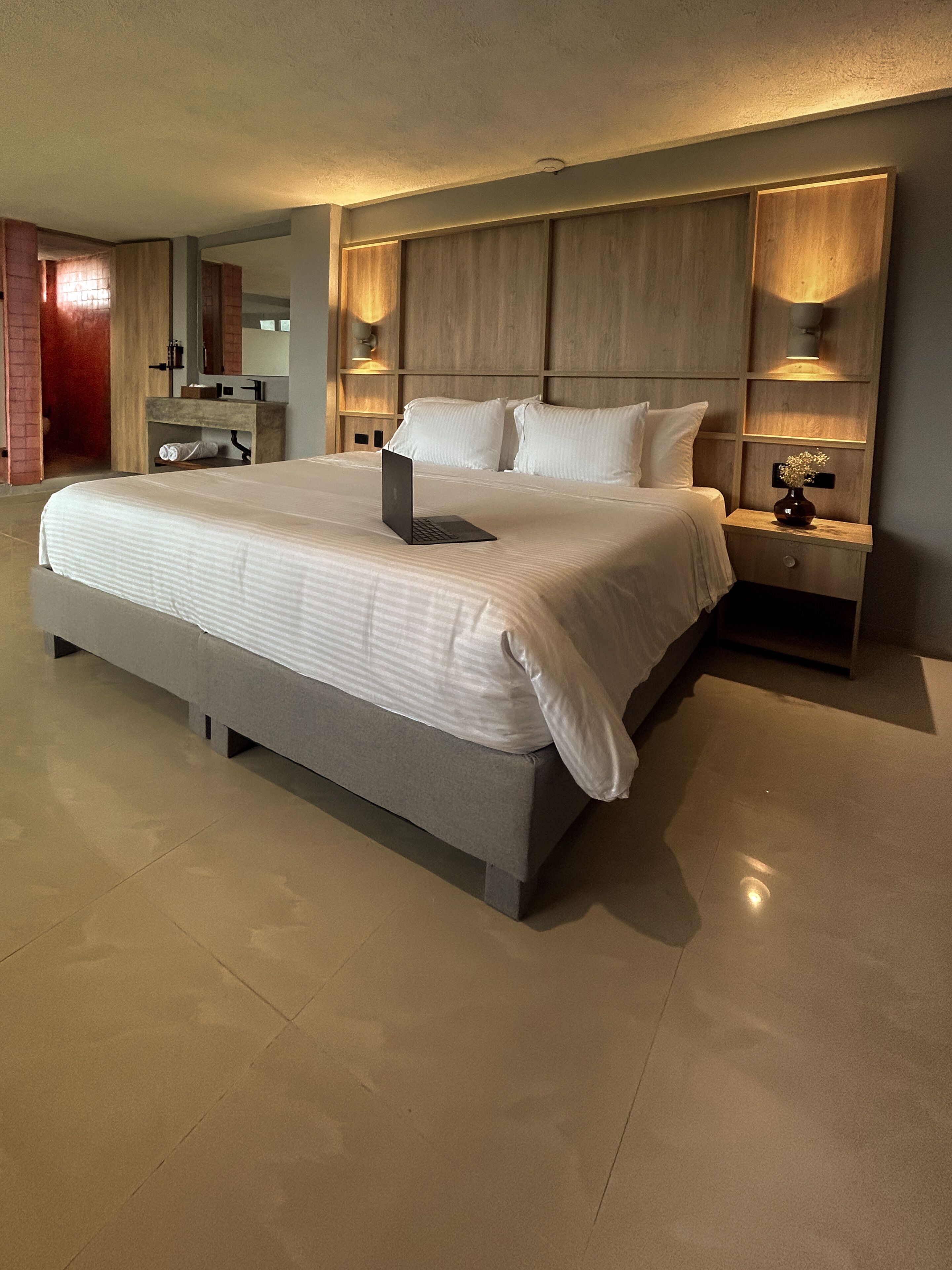 A modern hotel room with a large bed, a bedside table with a lamp, and a wooden headboard.