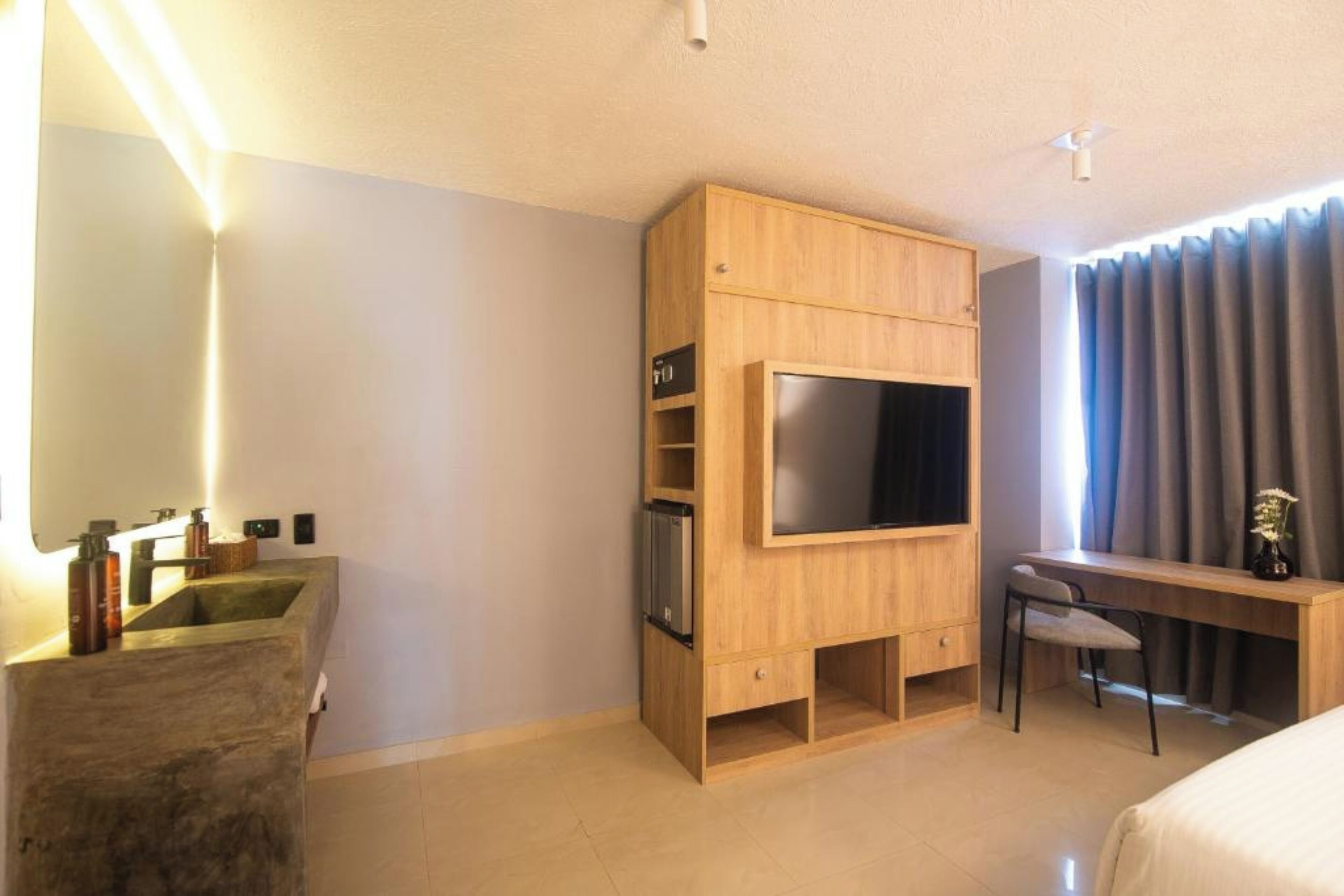 Quarto de hotel moderno com televisão integrada, escrivaninha com cadeira, janela grande com cortinas e lavatório de cimento.