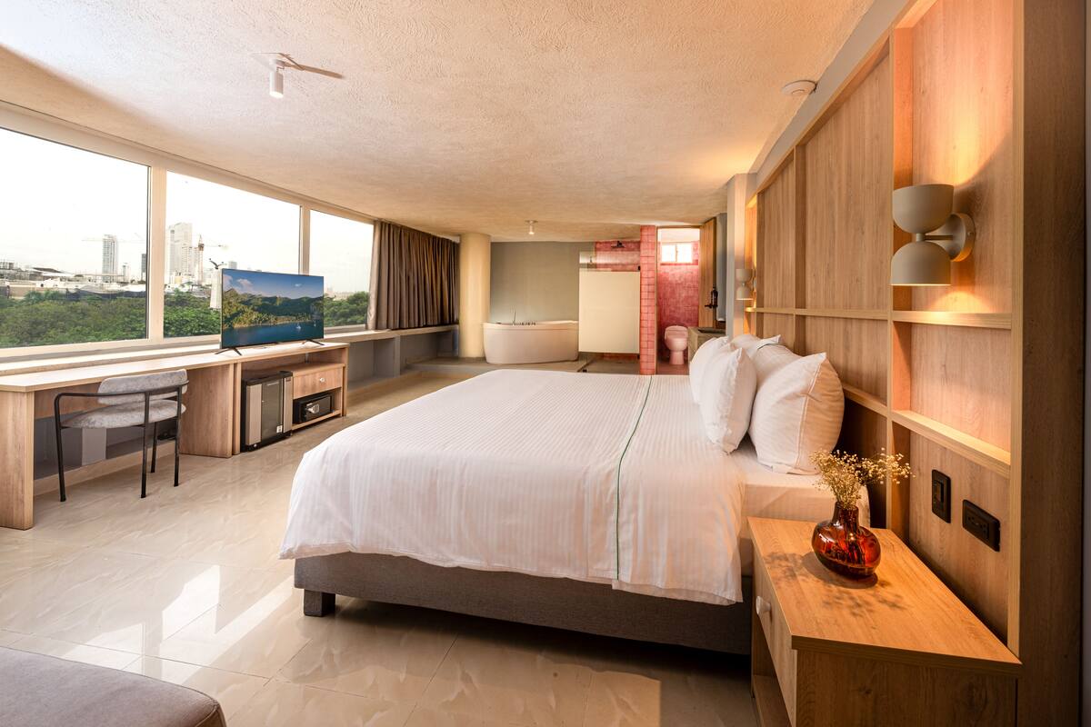 Quarto de hotel moderno com uma cama grande, uma mesa com computador, televisão e vista para o horizonte urbano.