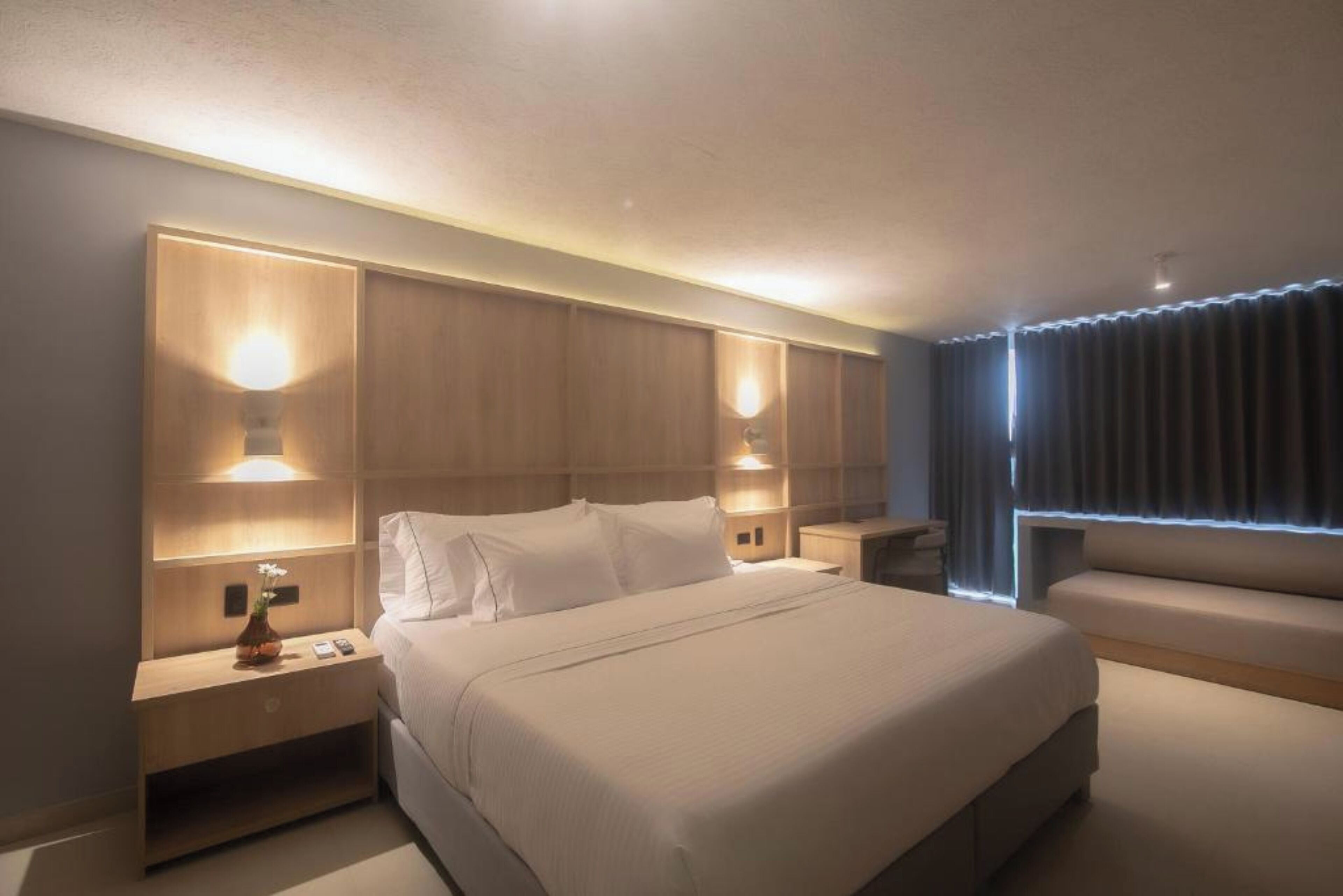 Ein modernes Hotelzimmer mit einem großen Bett, Nachttischen mit Lampen, einer Sitzecke und Blick auf die Stadt durch das Fenster.
