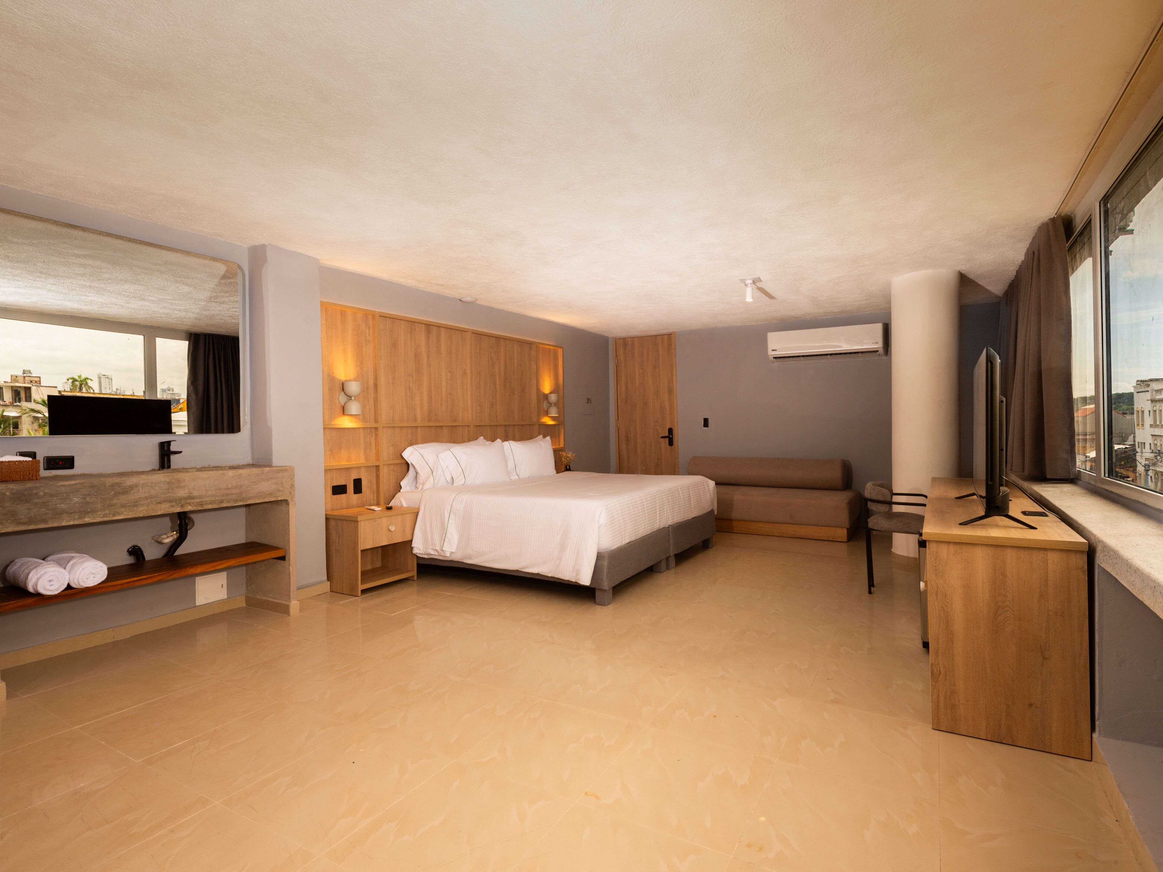 Una habitación de hotel moderna con una cama grande, un escritorio, una silla, un televisor y una ventana con vista a la ciudad.