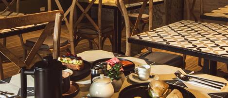 Comedor con sillas y mesas de madera, una cafetera, un hervidor de agua y un plato con sándwiches y fruta.