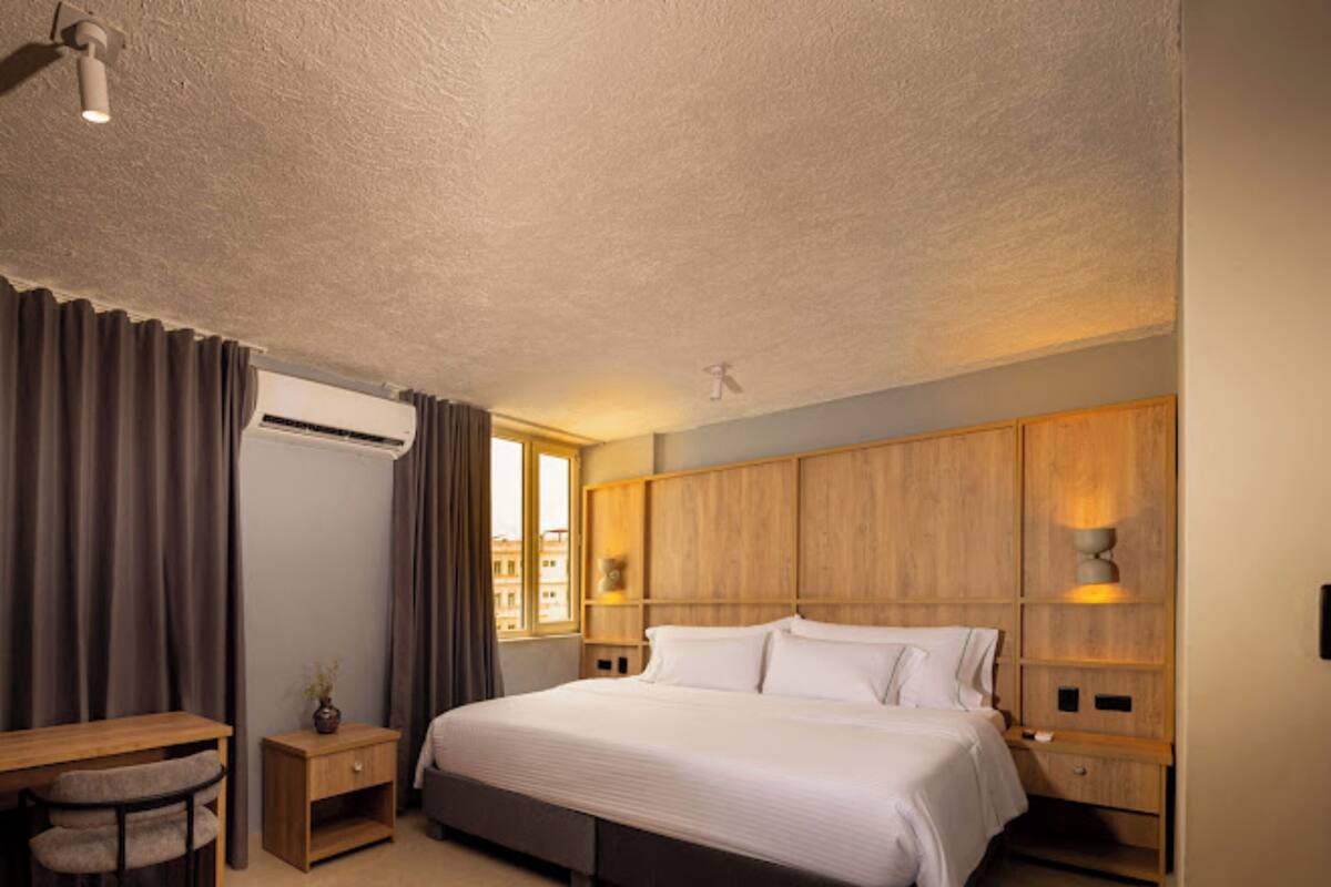 Quarto de hotel com cama, escrivaninha, cadeira, mesa de cabeceira, aparelho de ar condicionado e janela com cortinas.